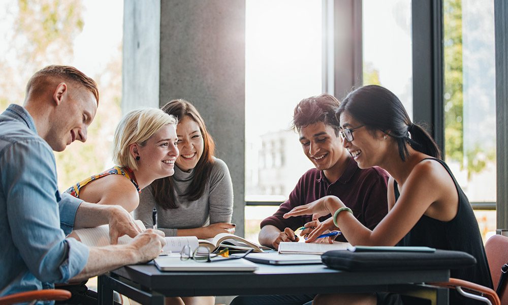 Blog - College Students Sitting at a Table Talking and Laughing Next to a Large Window with Sunshine Pouring Through